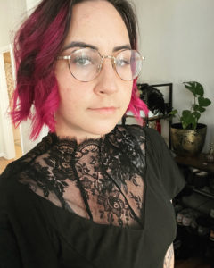 Self portrait of Erin Gordon - a tan woman with pink, chin length curled hair, wearing a black lace dress and round glasses